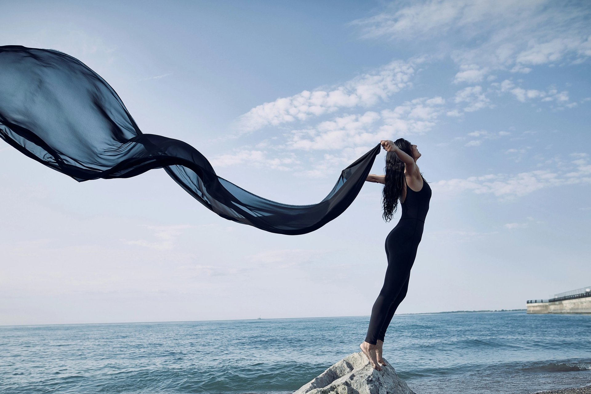 Frau steht mit wehendem Seidenschal am Meer.