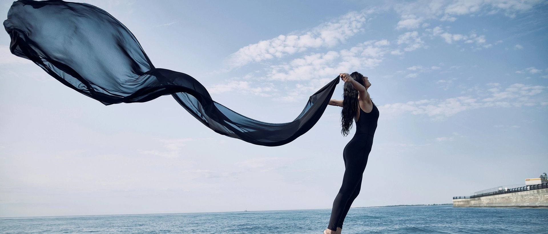 Frau steht mit wehendem Seidenschal am Meer.