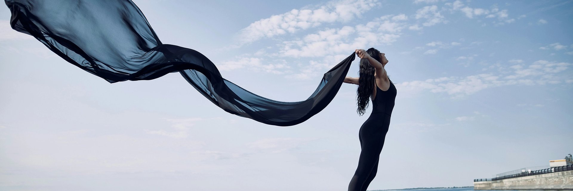 Frau steht mit wehendem Seidenschal am Meer.