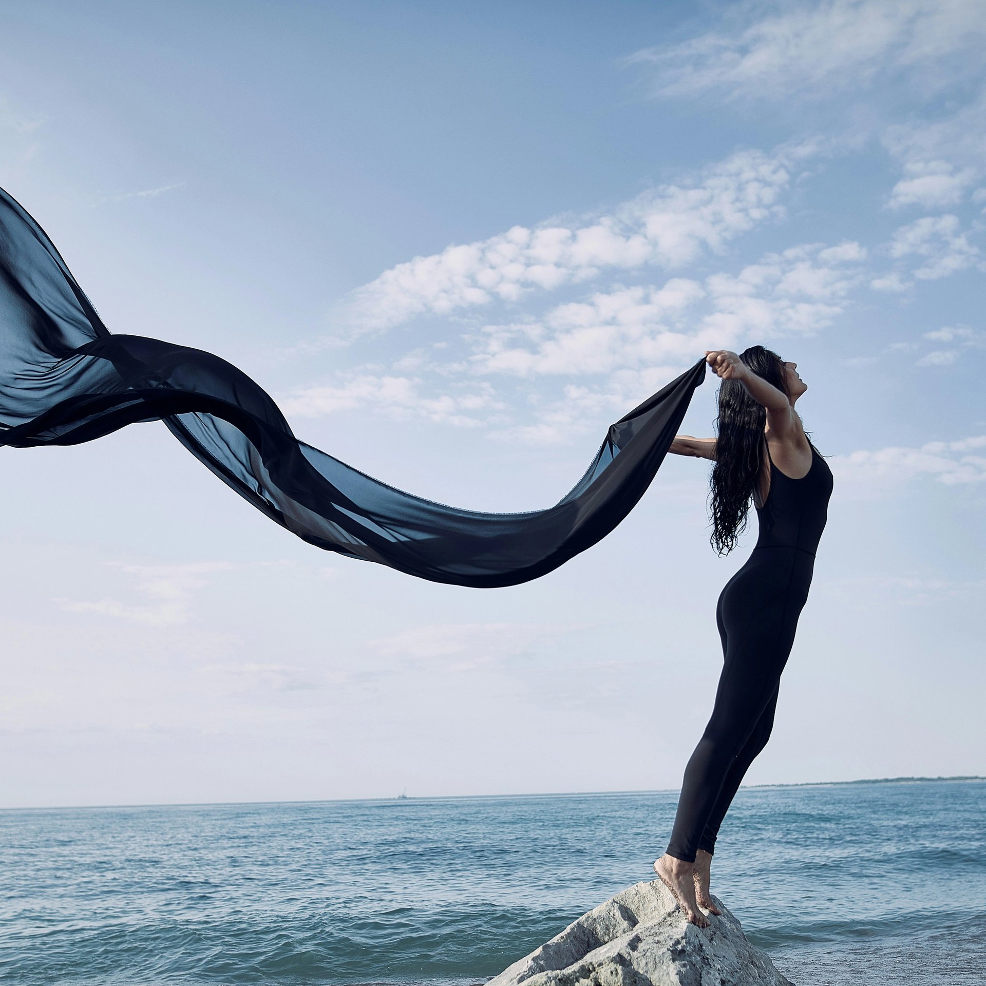 Frau steht mit wehendem Seidenschal am Meer.