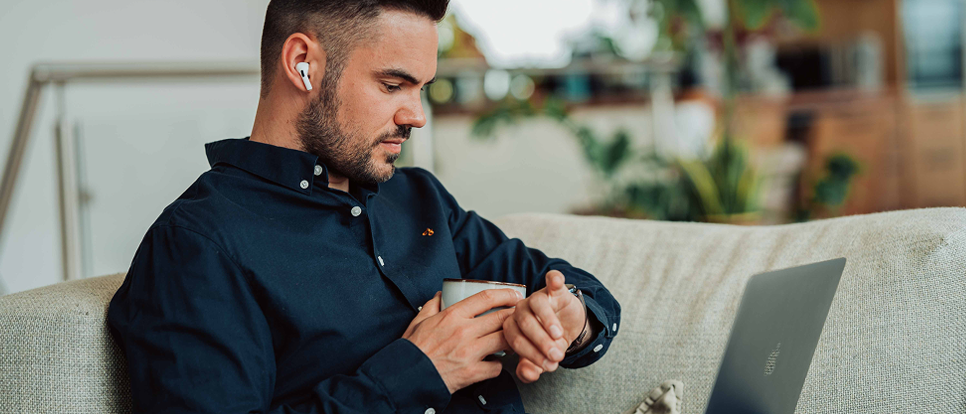 A man is sitting on the couch with his laptop and looking at his watch.