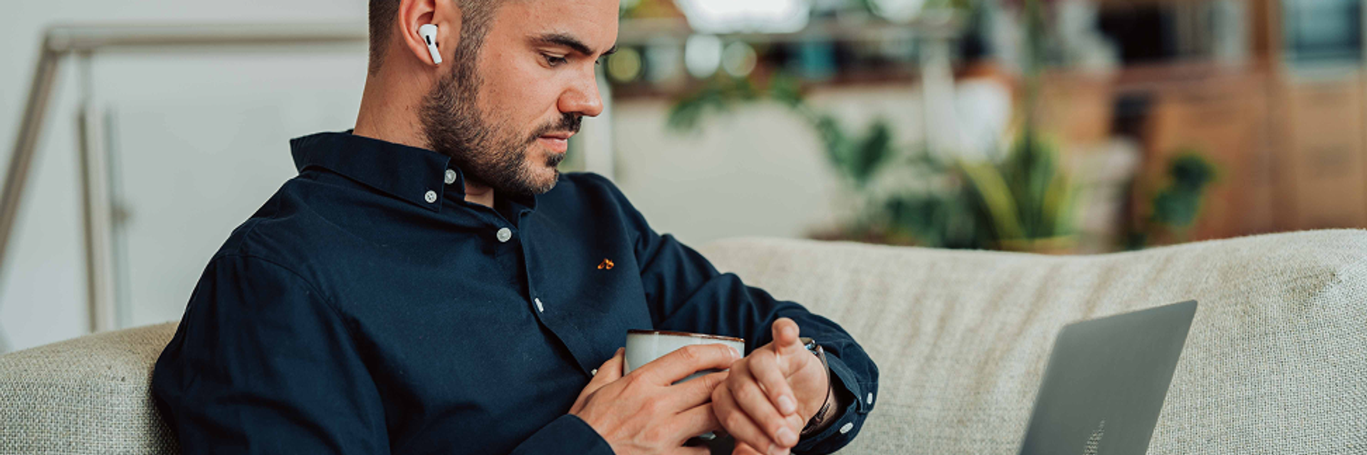 Ein Mann sitzt mit Laptop auf der der Couch und schaut auf seine Uhr.