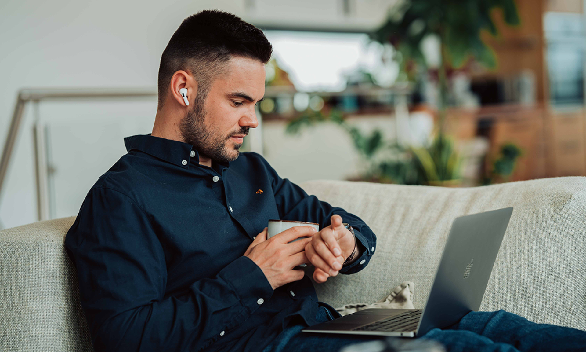 A man is sitting on the couch with his laptop and looking at his watch.