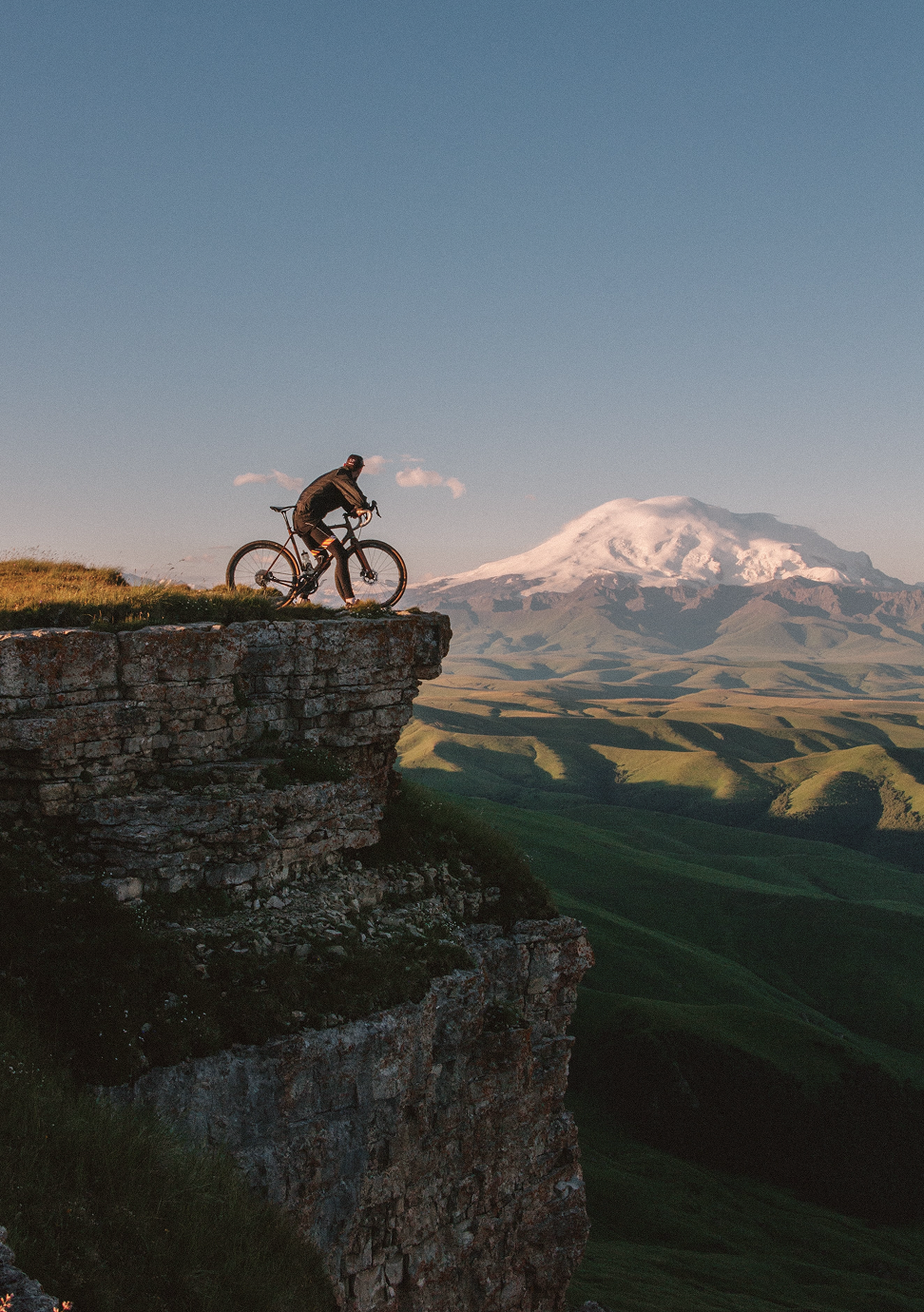 Ein Fahrradfahrer steht an einer Felskante, im Hintergrund ein schneebedeckter Berg.