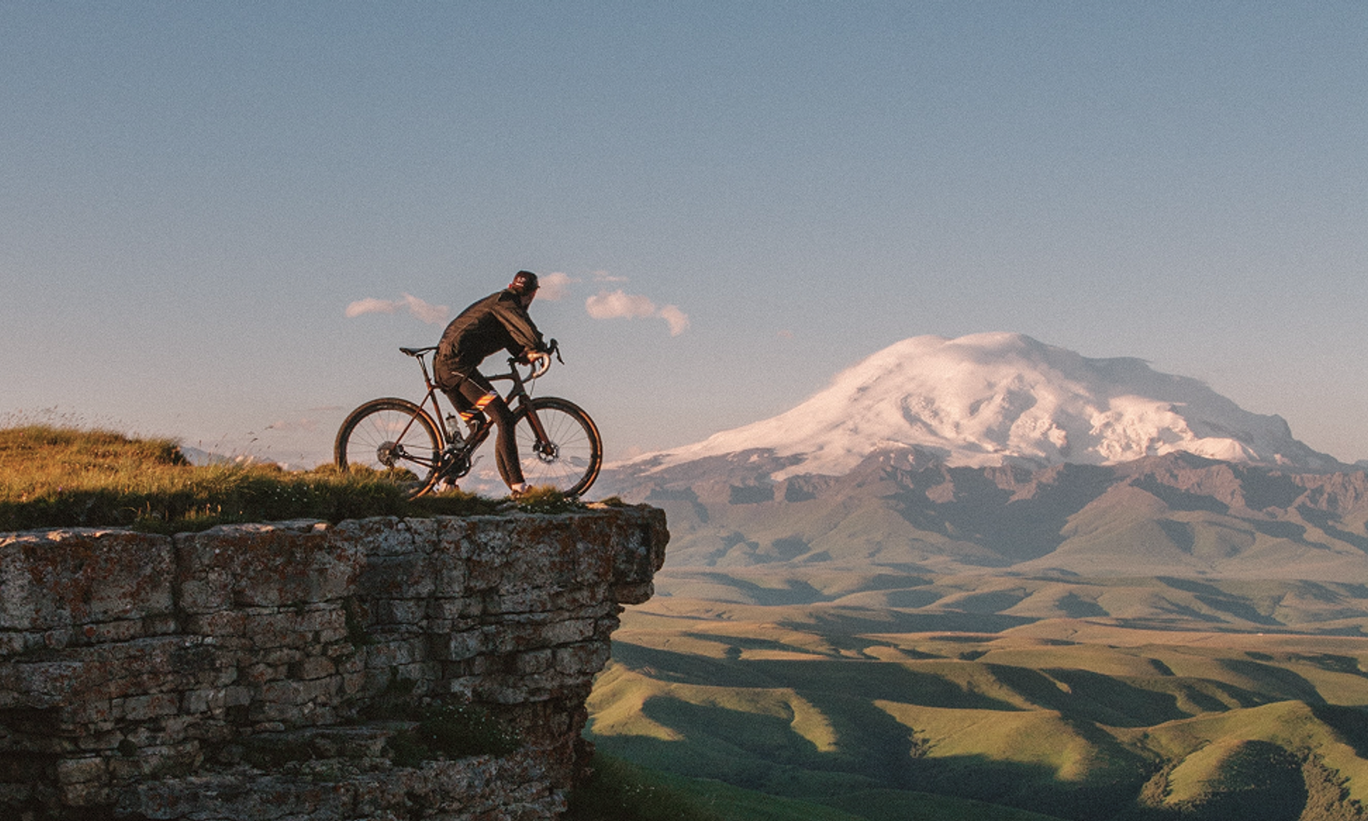 Ein Fahrradfahrer steht an einer Felskante, im Hintergrund ein schneebedeckter Berg.