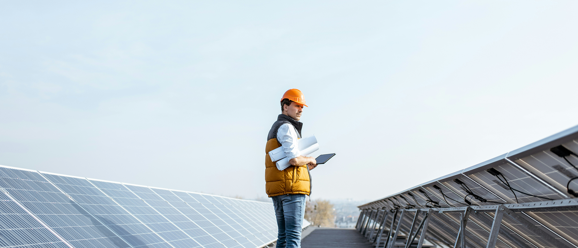 Ingenieur steht mit Tablet und Salesforce Energy & Utilities Cloud und aufgerollten Plänen zwischen Solarpanelen.