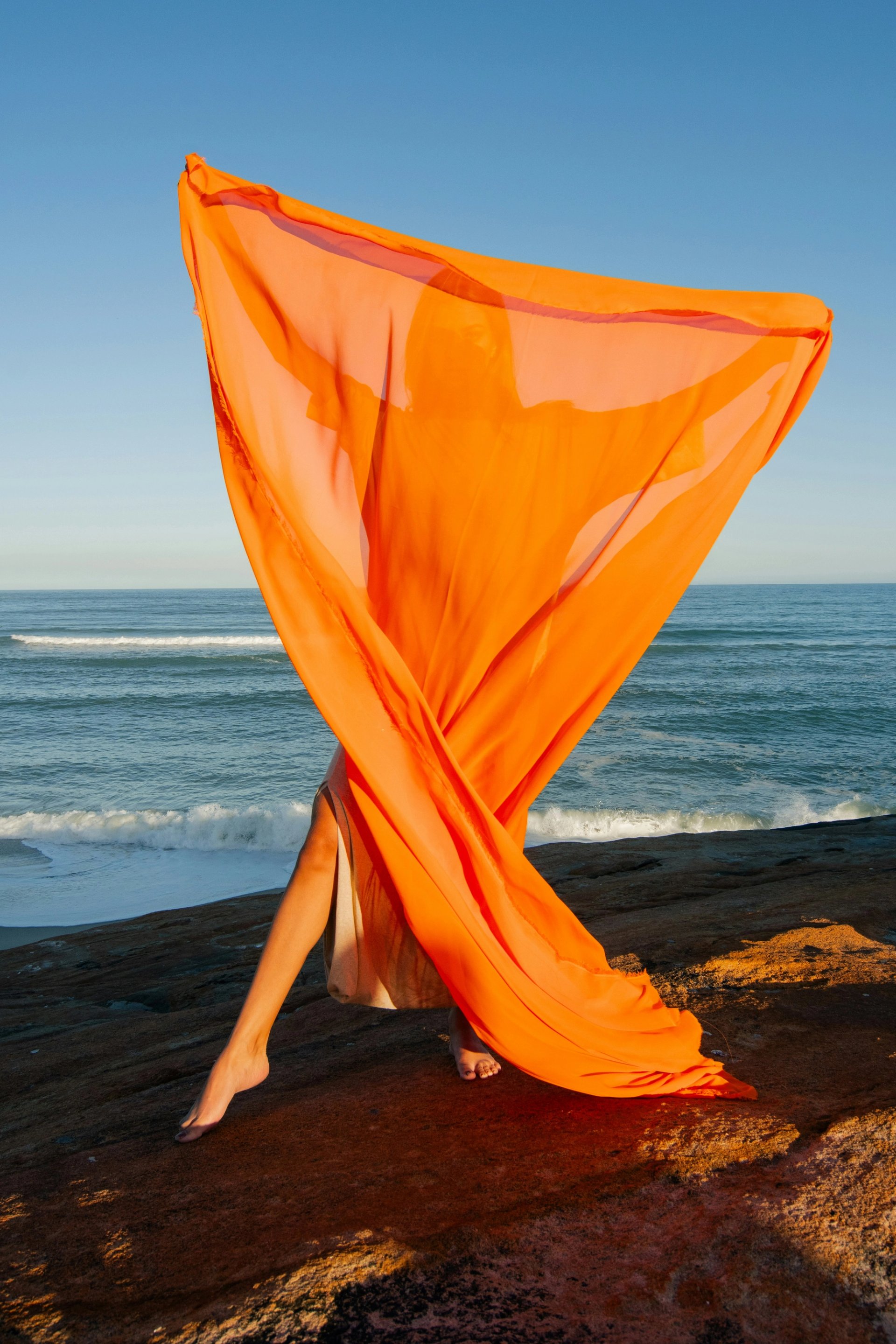 Frau steht am Meer und hält ein orangenes Tuch vor sich.