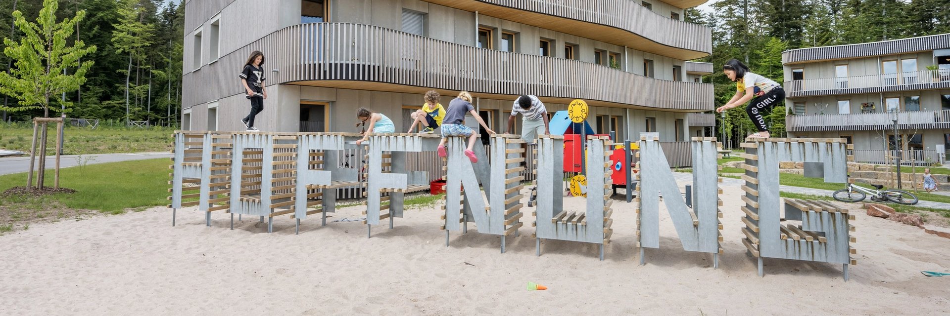 Kinder spielen auf einem Hoffnung-Schriftzug vor einem Holzhaus.