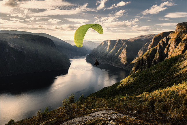 A yellow paraglider flies over a river valley lined with steep mountains.