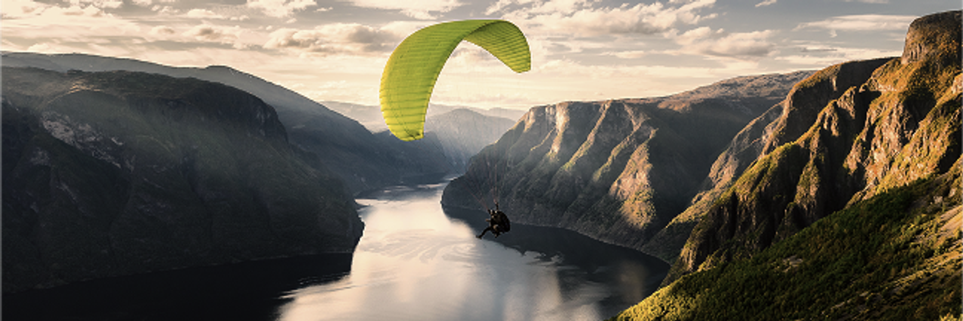 A yellow paraglider flies over a river valley lined with steep mountains.