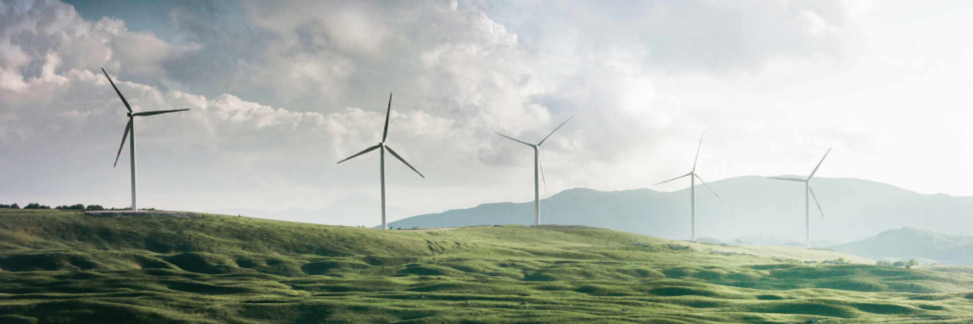 Fünf Windräder stehen auf einem grünen Hügel vor wolkigem Himmel.