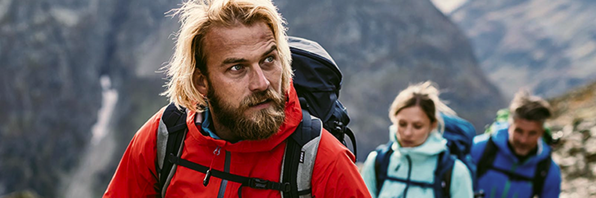 Ein Mann mit roter Schöffel-Jacke wandert in den Bergen, hinter ihm eine Frau und ein Mann.
