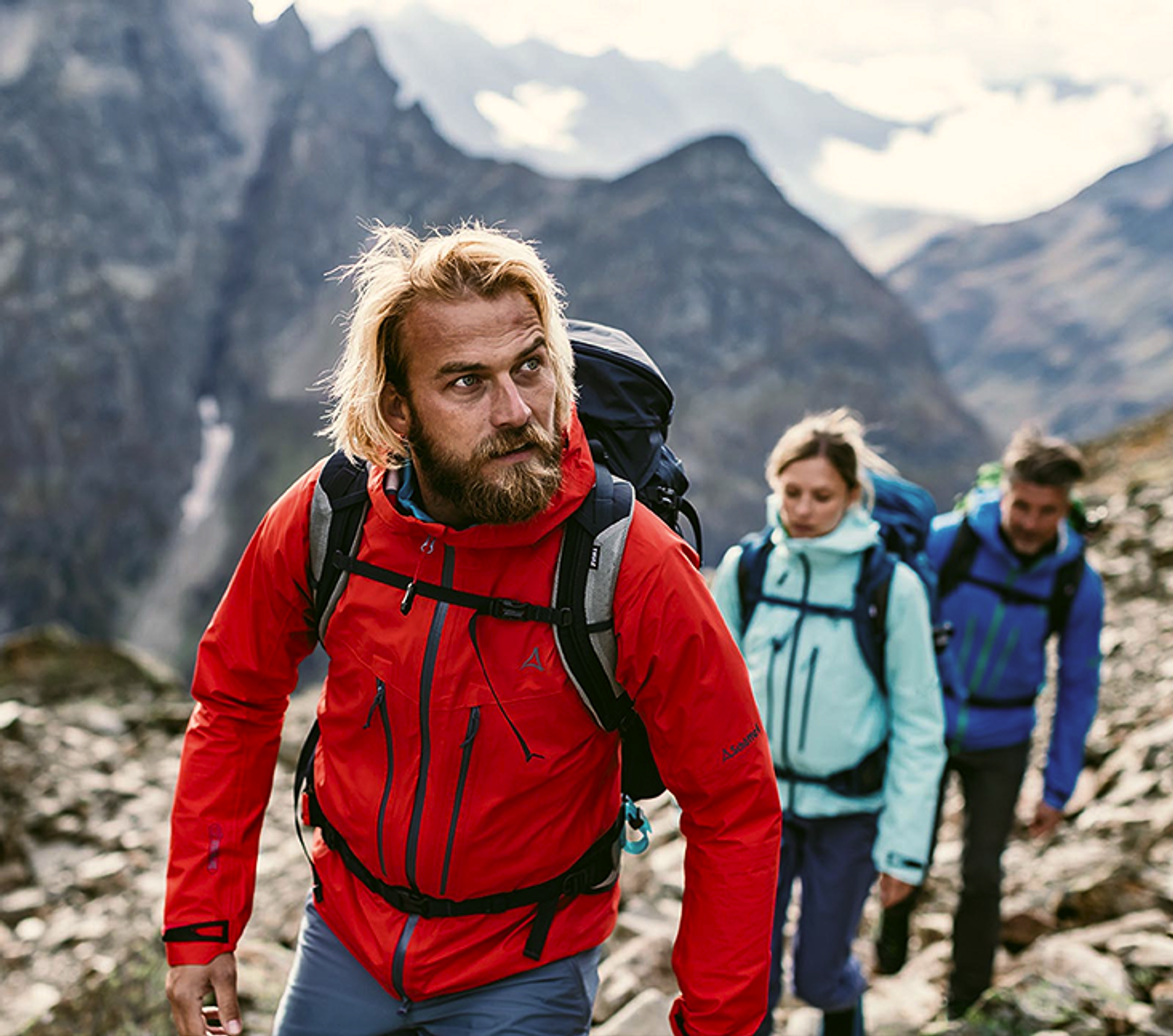 Ein Mann mit roter Schöffel-Jacke wandert in den Bergen, hinter ihm eine Frau und ein Mann.