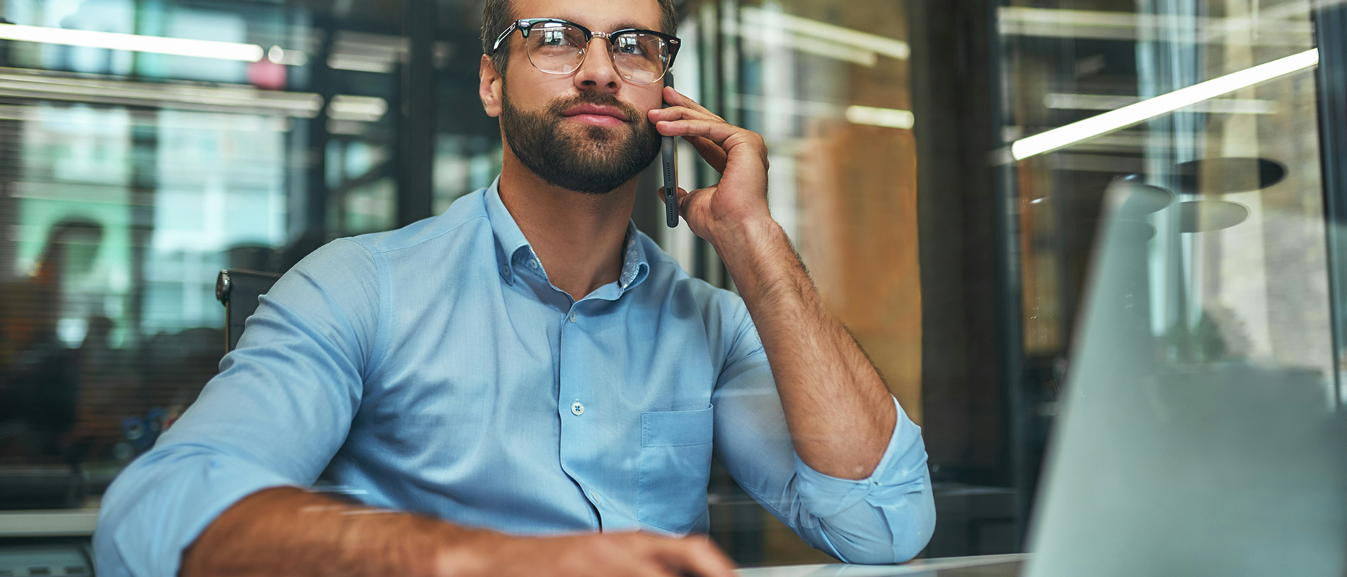 Mann sitzt am Schreibtisch vor Laptop mit Service Cloud und telefoniert mit Handy.