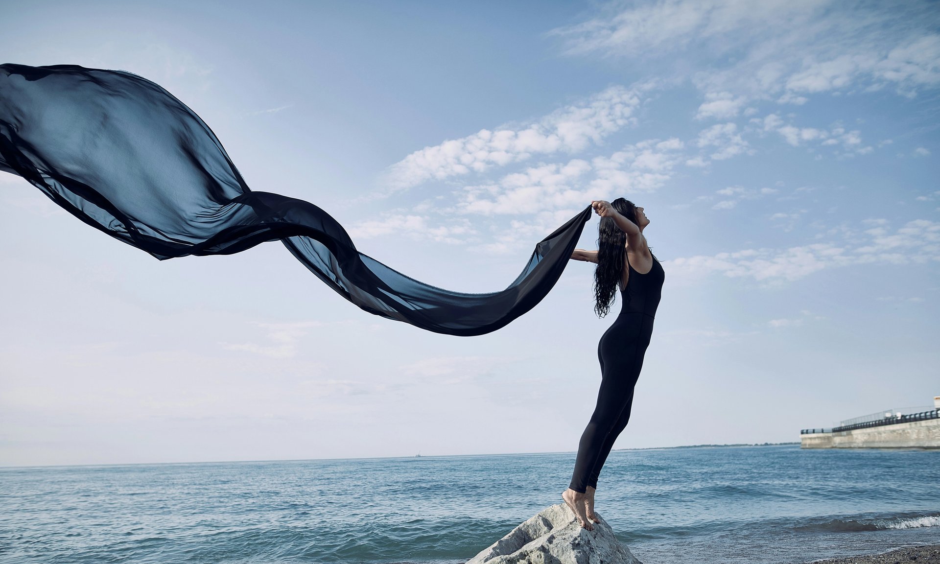 Frau steht mit wehendem Seidenschal am Meer.