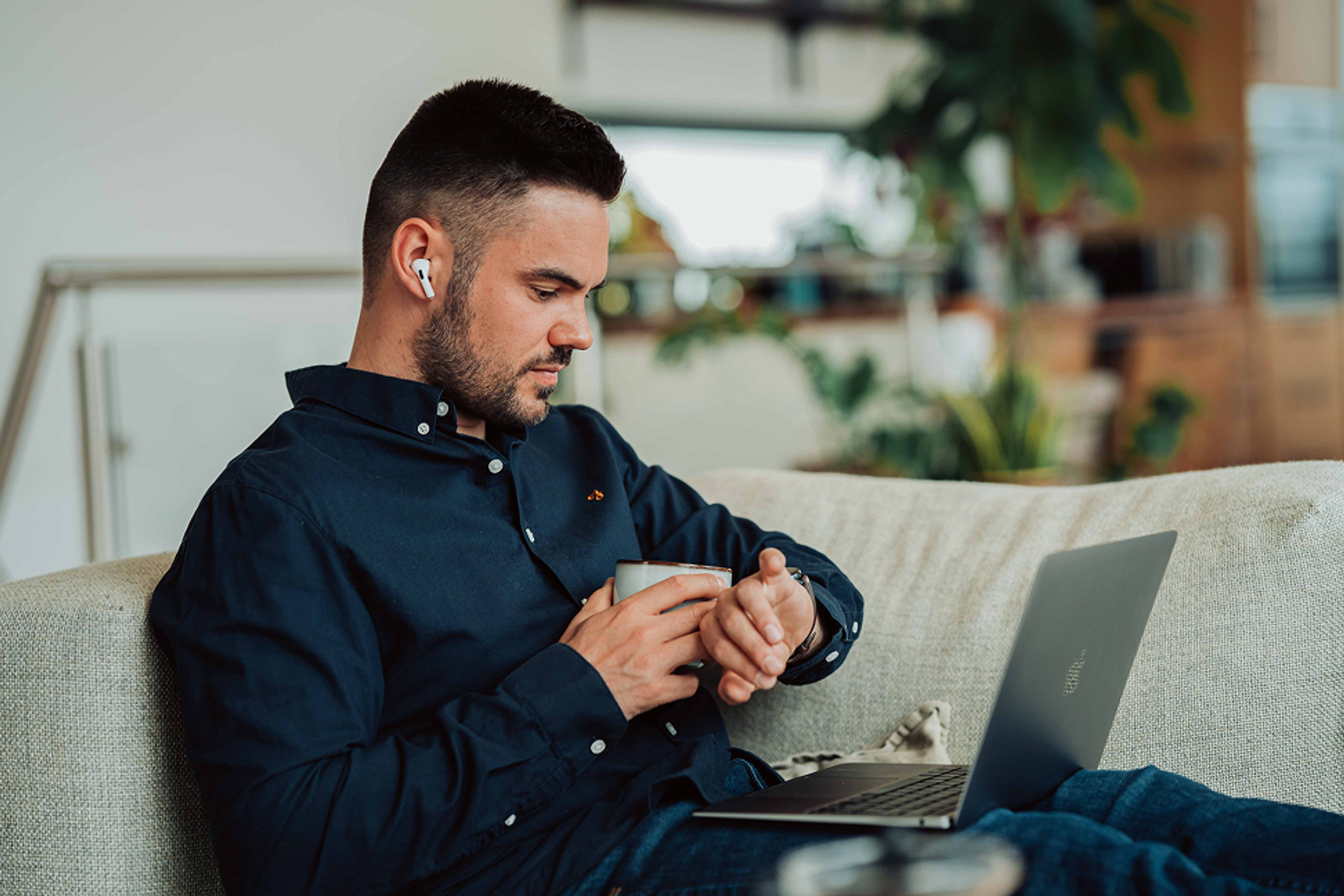 Ein Mann sitzt mit Laptop auf der der Couch und schaut auf seine Uhr. 