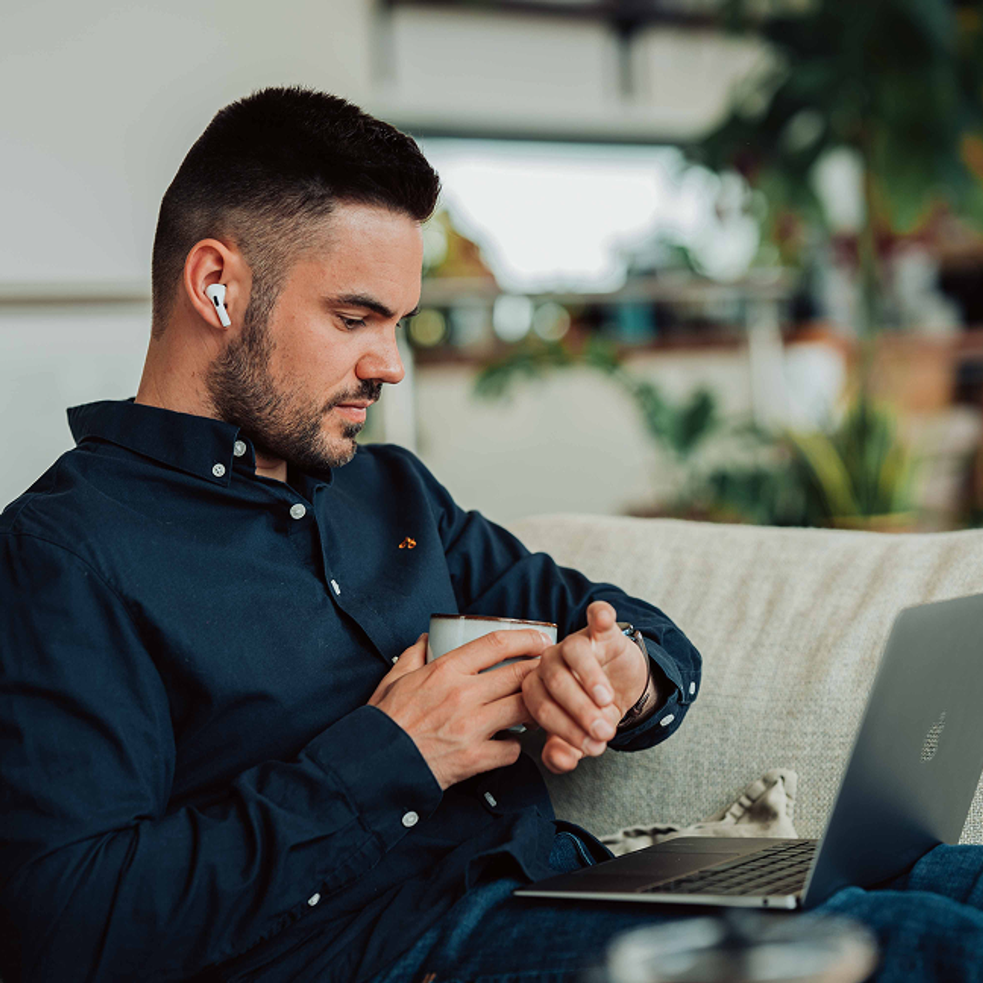 Ein Mann sitzt mit Laptop auf der der Couch und schaut auf seine Uhr.