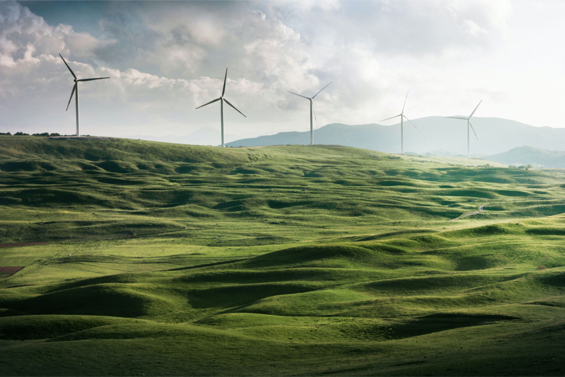 Fünf Windräder stehen auf einem grünen Hügel vor wolkigem Himmel.