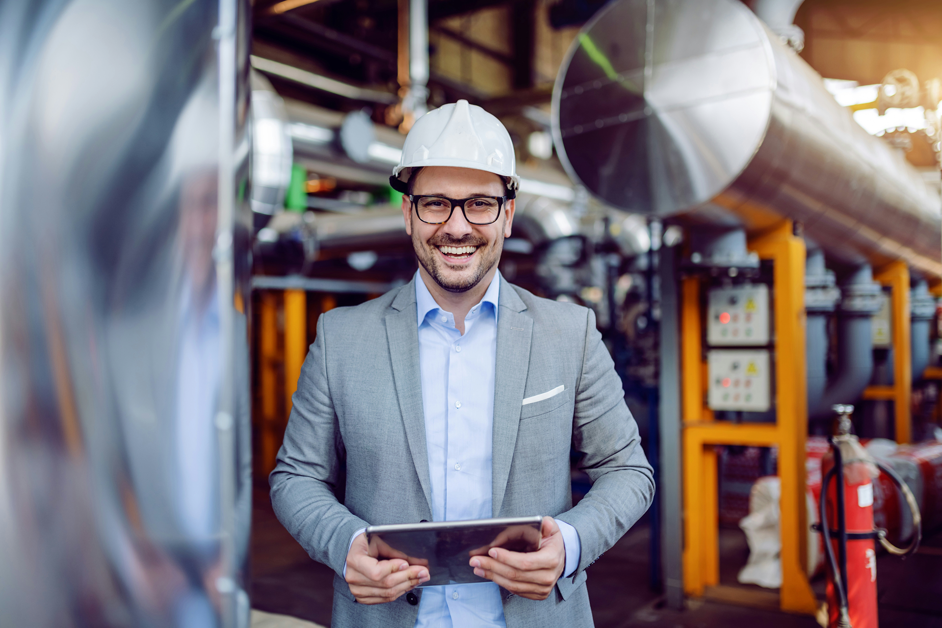 Mann mit Helm und Tablet mit Manufacturing Cloud steht in Fertigungshalle und schaut in Kamera.