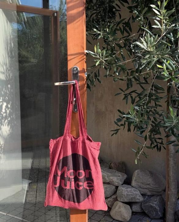 A red "Moon Juice" tote bag hangs from a door handle outdoors, beside an olive tree.