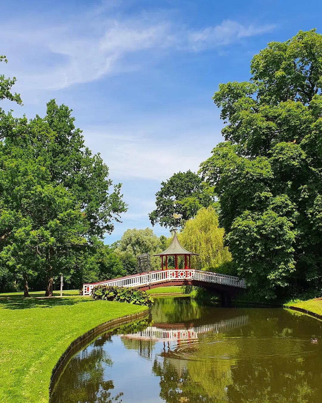 An ornate red and white bridge with a central pagoda-style pavilion spans a tranquil stream in a lush green park under a blue sky.