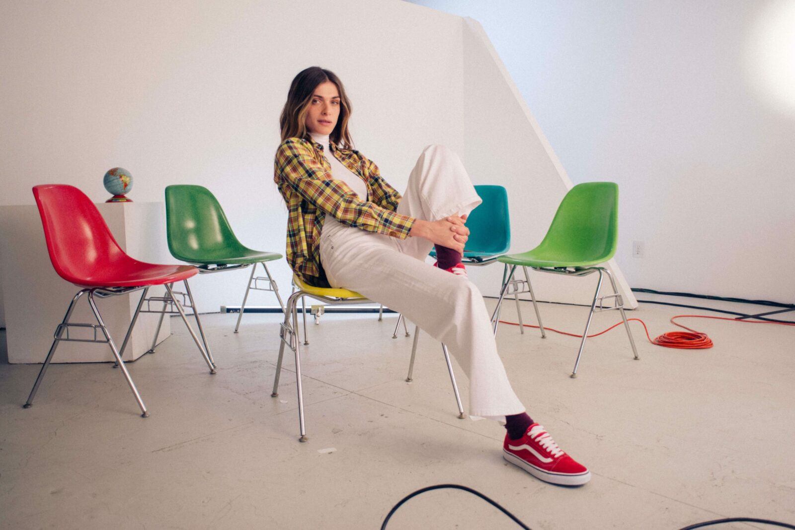 A woman in a white turtleneck, plaid shirt, and white wide-leg pants sits on a yellow chair, holding her leg.