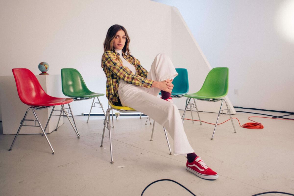 A woman in a white turtleneck, plaid shirt, and white wide-leg pants sits on a yellow chair, holding her leg.