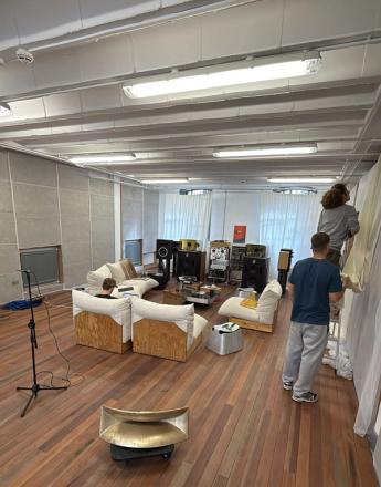 A spacious room with wooden floors and exposed beams, set up with high-end audio equipment and modern white seating. Two people hang white fabric on a wall, while another sits on a sofa with a laptop. A large golden horn speaker is in the foreground.