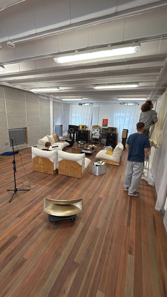 A spacious room with hardwood floors features a large sound system, white couches, and two men setting up white drapes, while a gold horn speaker sits in the foreground.