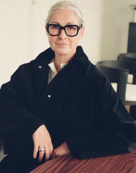 A woman with gray hair and thick black glasses, wearing a black jacket, sits at a wooden table and looks at the camera.
