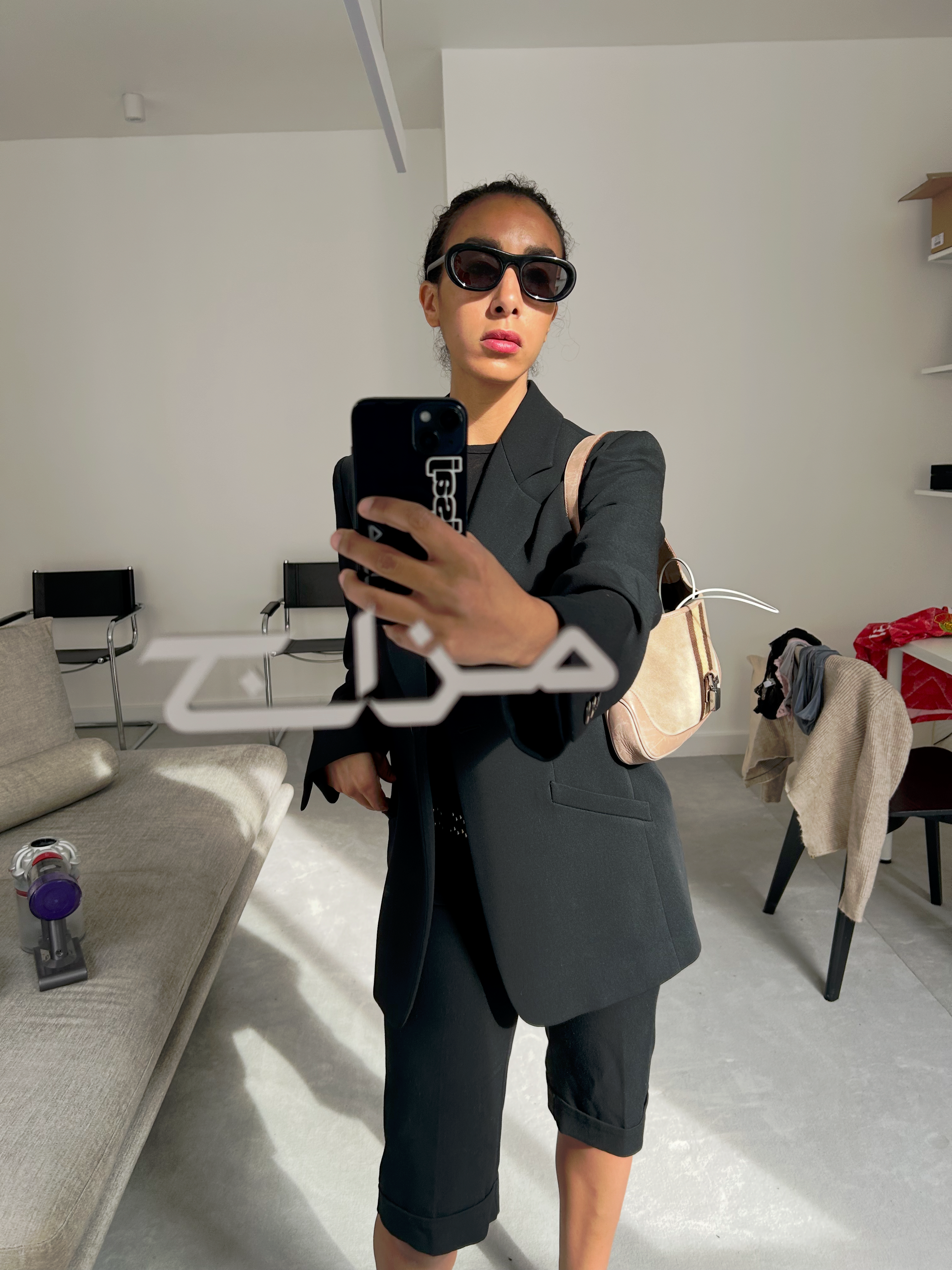 A person in oversized black sunglasses, a black blazer, and shorts takes a mirror selfie with their phone in a bright, minimalist room.