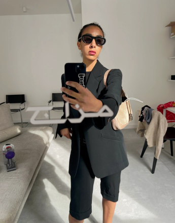 A person in oversized black sunglasses, a black blazer, and shorts takes a mirror selfie with their phone in a bright, minimalist room.