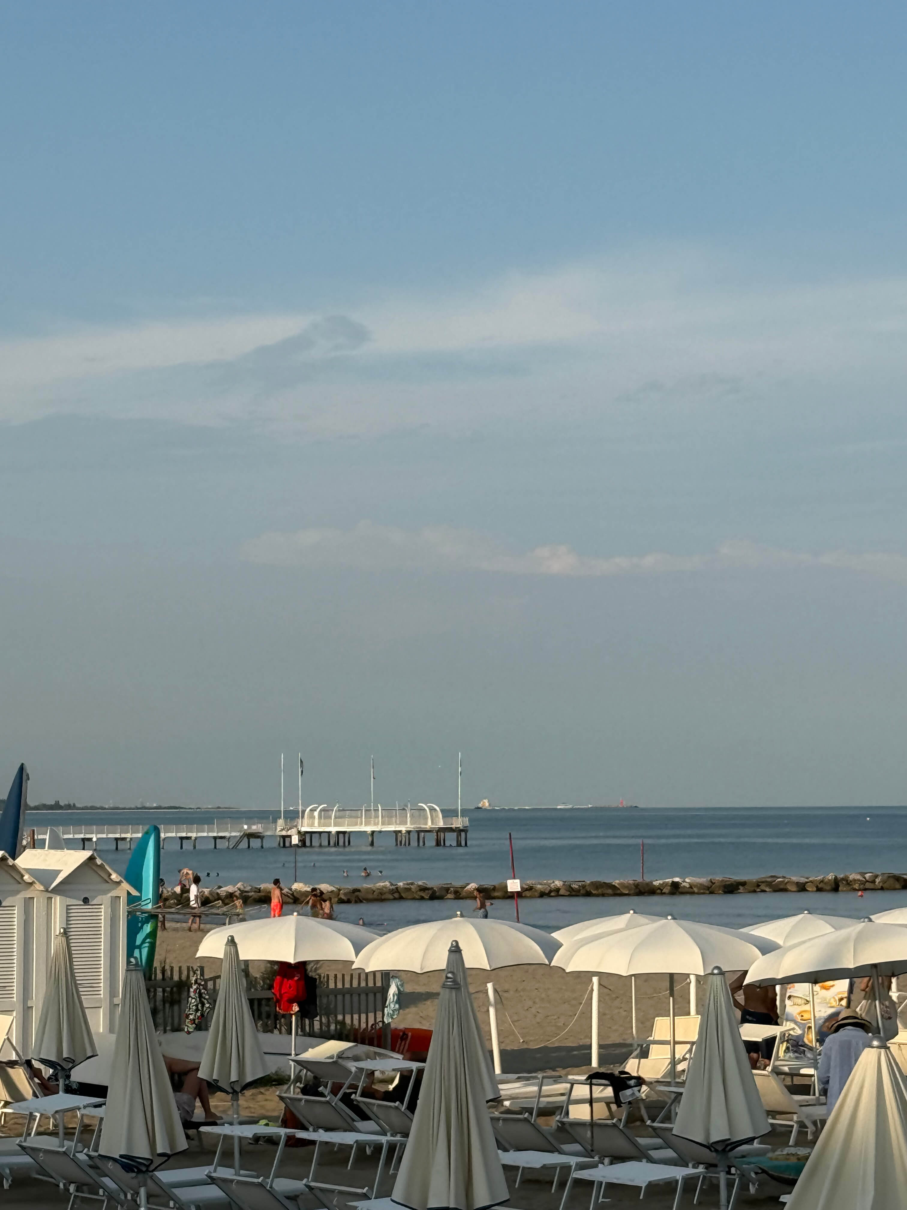 A sunny beach with white umbrellas and lounge chairs, people by the water, and a pier extending into the sea under a blue sky.