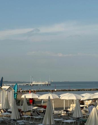 A sunny beach with white umbrellas and lounge chairs, people by the water, and a pier extending into the sea under a blue sky.