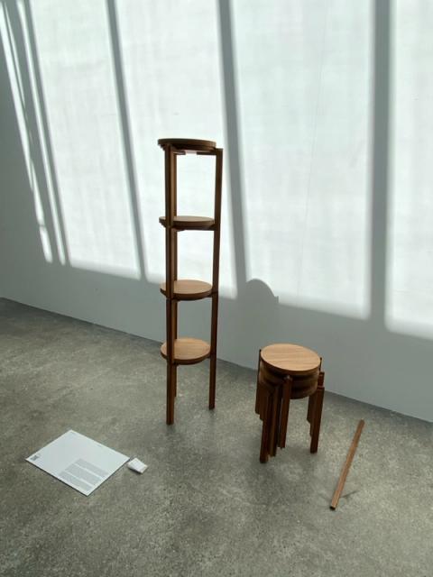 A tall wooden shelf unit and three stacked wooden stools on a concrete floor with strong window shadows.
