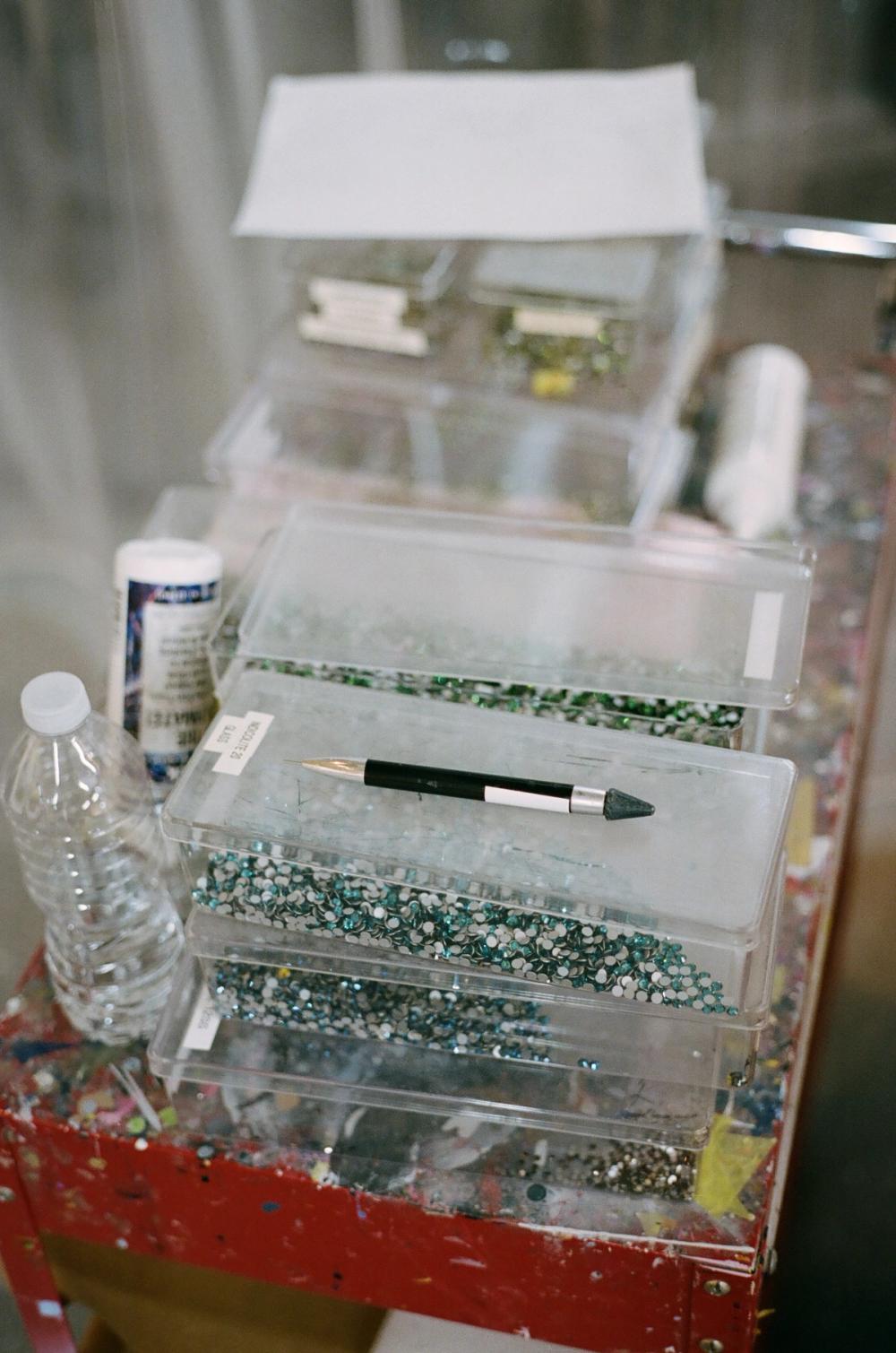 A stack of clear containers filled with rhinestones on a paint-splattered red table, with a rhinestone picker tool.