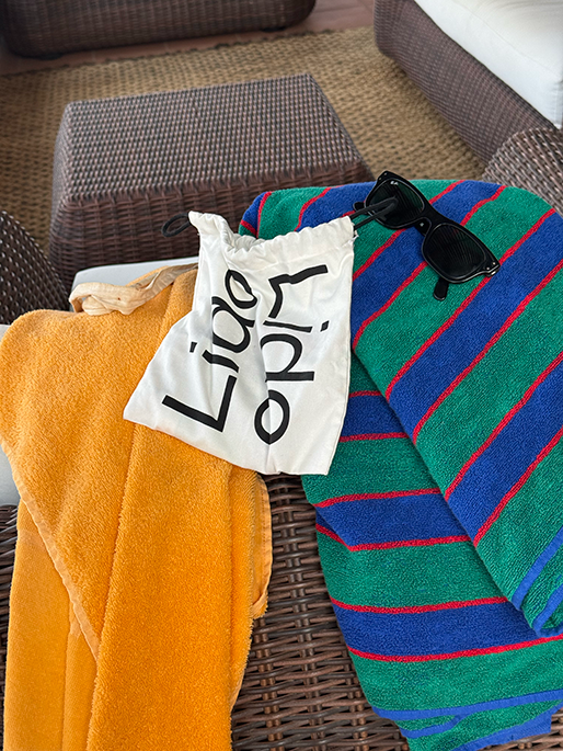 Orange towel, striped towel, white Lido bag, and black sunglasses on a wicker lounge chair.