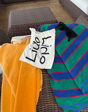 Orange towel, striped towel, white Lido bag, and black sunglasses on a wicker lounge chair.