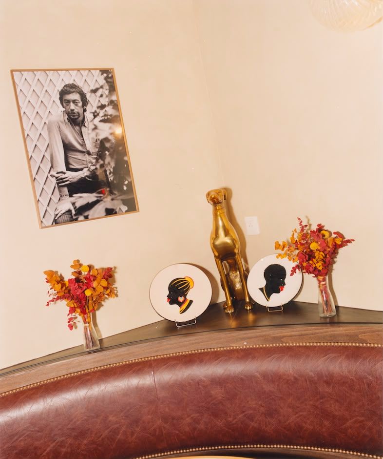 A framed photo of Serge Gainsbourg hangs above a golden dog statue, two silhouette plates, and dried floral arrangements on a shelf, with a leather booth in the foreground.