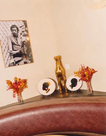 A framed photo of Serge Gainsbourg hangs above a golden dog statue, two silhouette plates, and dried floral arrangements on a shelf, with a leather booth in the foreground.