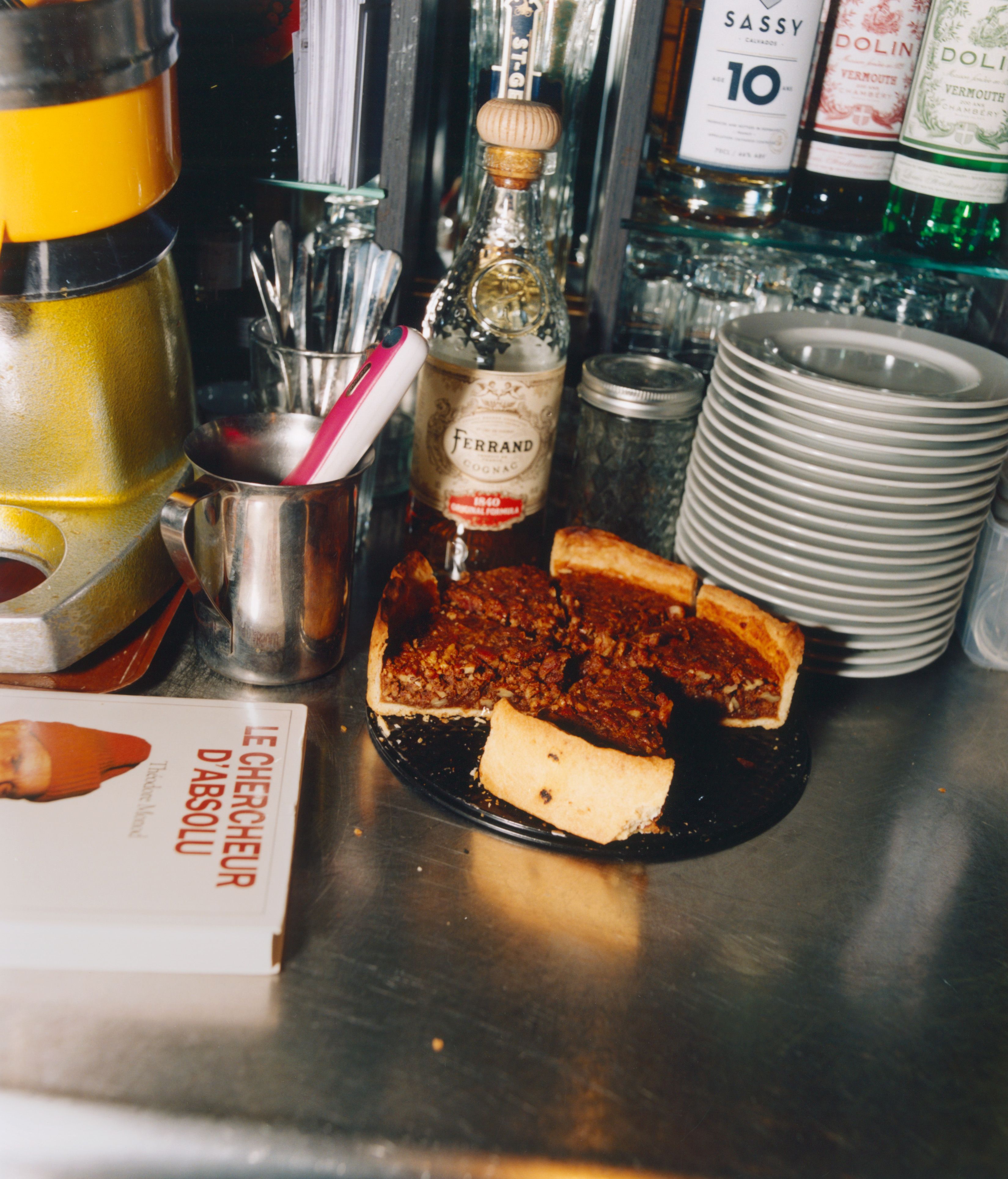 A pecan pie, a bottle of Ferrand cognac, stacked plates, and a book on a bar counter.