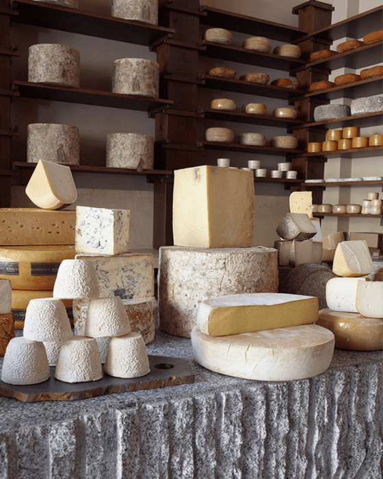 A large variety of cheeses displayed on a carved stone table and wooden shelves.