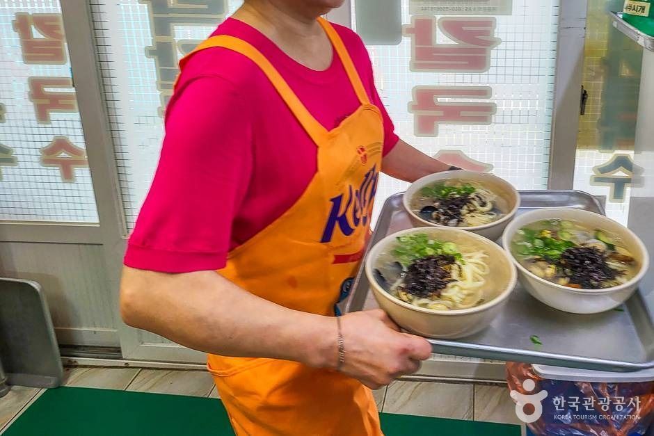 A person in a pink shirt and orange apron carries a tray with three bowls of noodle soup topped with seaweed and green onions.