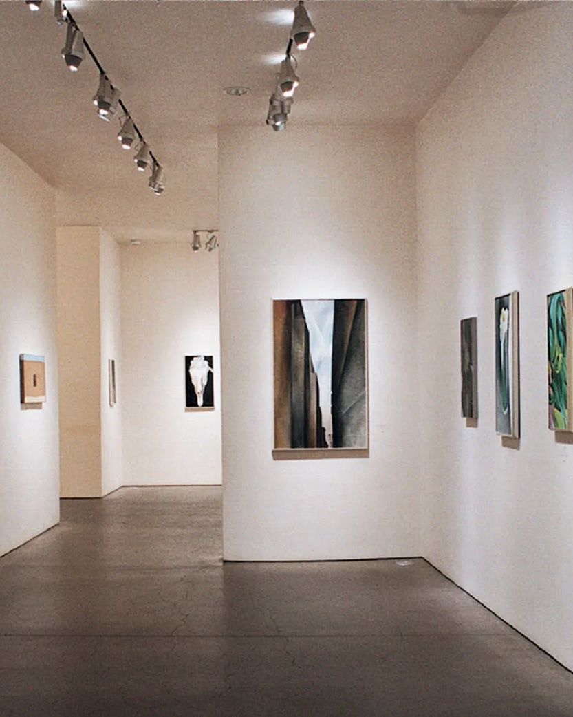 A brightly lit art gallery with white walls, a dark floor, and several paintings under track lighting.