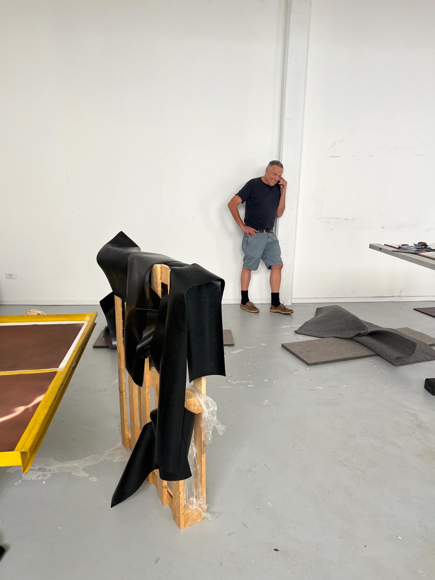 A man talks on the phone in a large, white room with black fabric draped over a wooden pallet, gray felt pieces on the floor, and a yellow-framed panel.