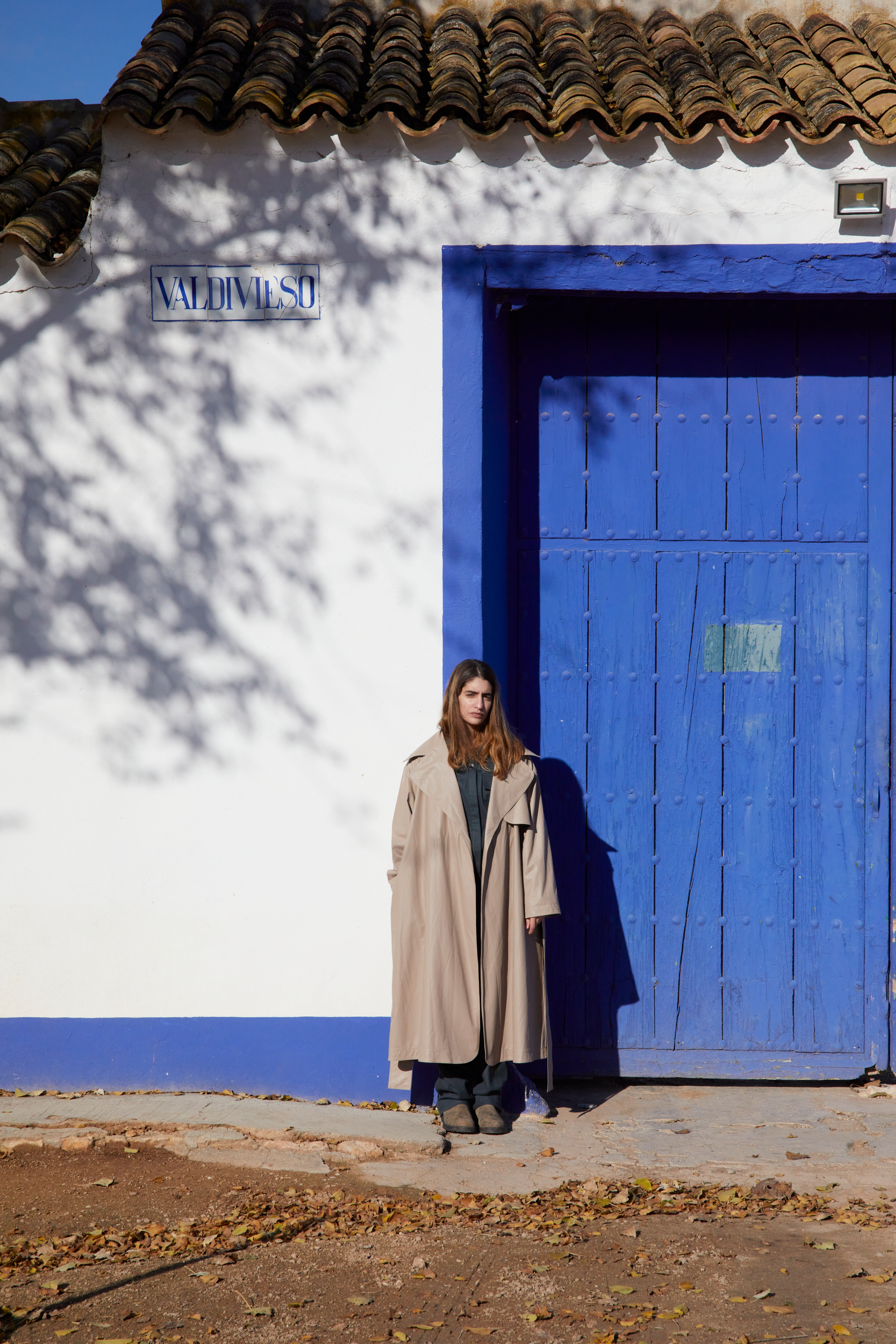 A woman in a long beige coat stands against a white wall and a vibrant blue door, with tree shadows on the wall.