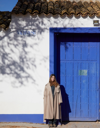 A woman in a long beige coat stands against a white wall and a vibrant blue door, with tree shadows on the wall.