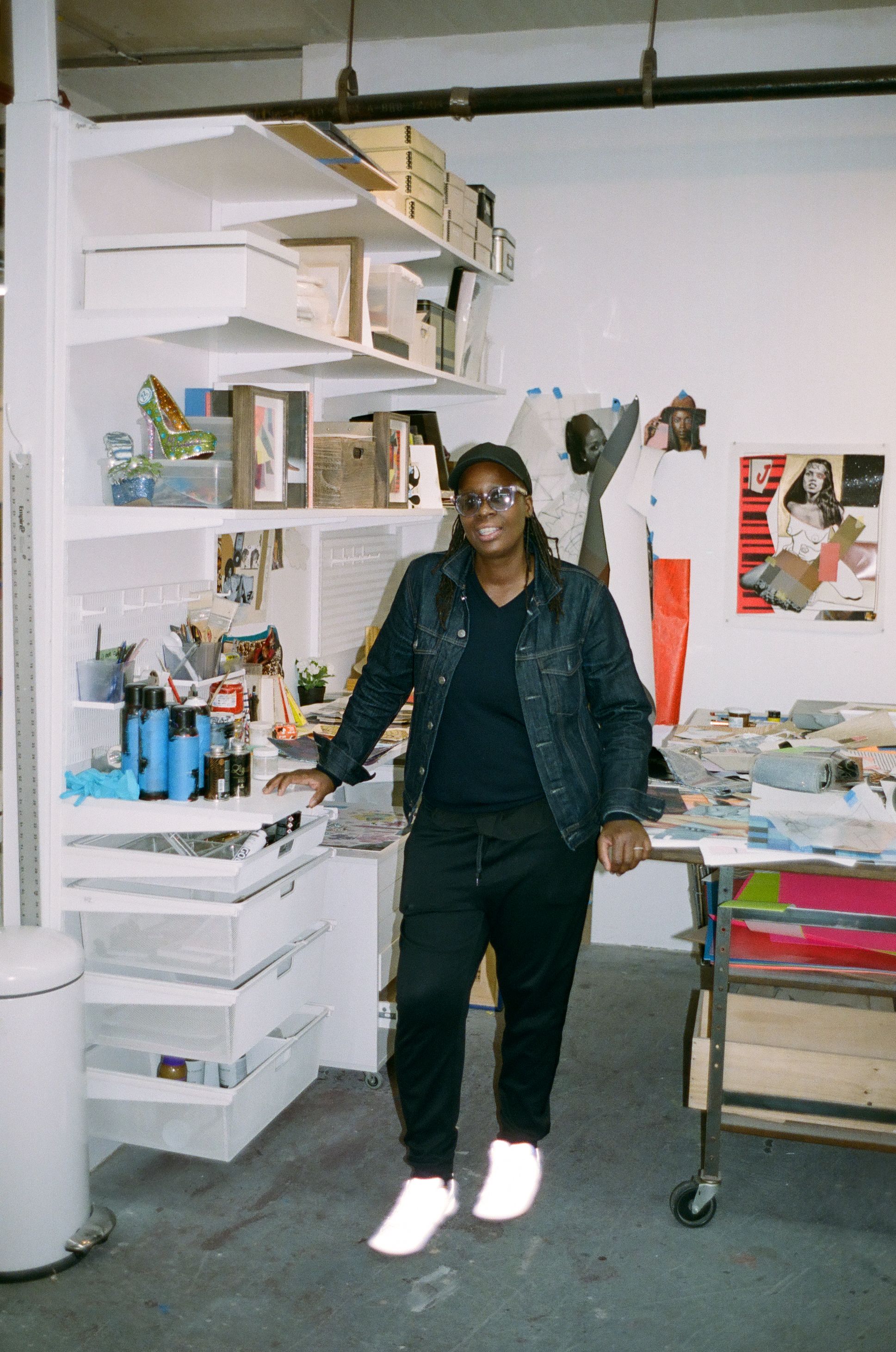 A smiling person in a denim jacket, black pants, and glowing white sneakers stands in a busy art studio.