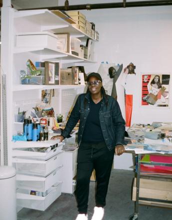 A smiling person in a denim jacket, black pants, and glowing white sneakers stands in a busy art studio.
