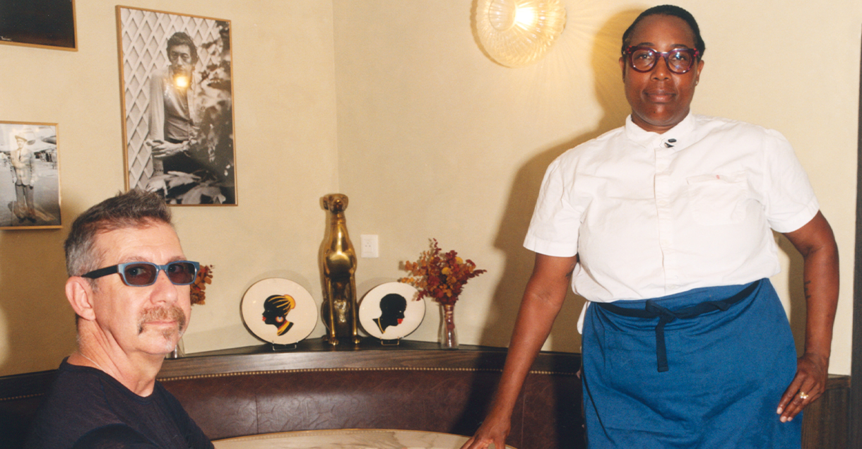 A man in sunglasses sits at a table while a woman in a chef's uniform stands beside him in a decorated room.